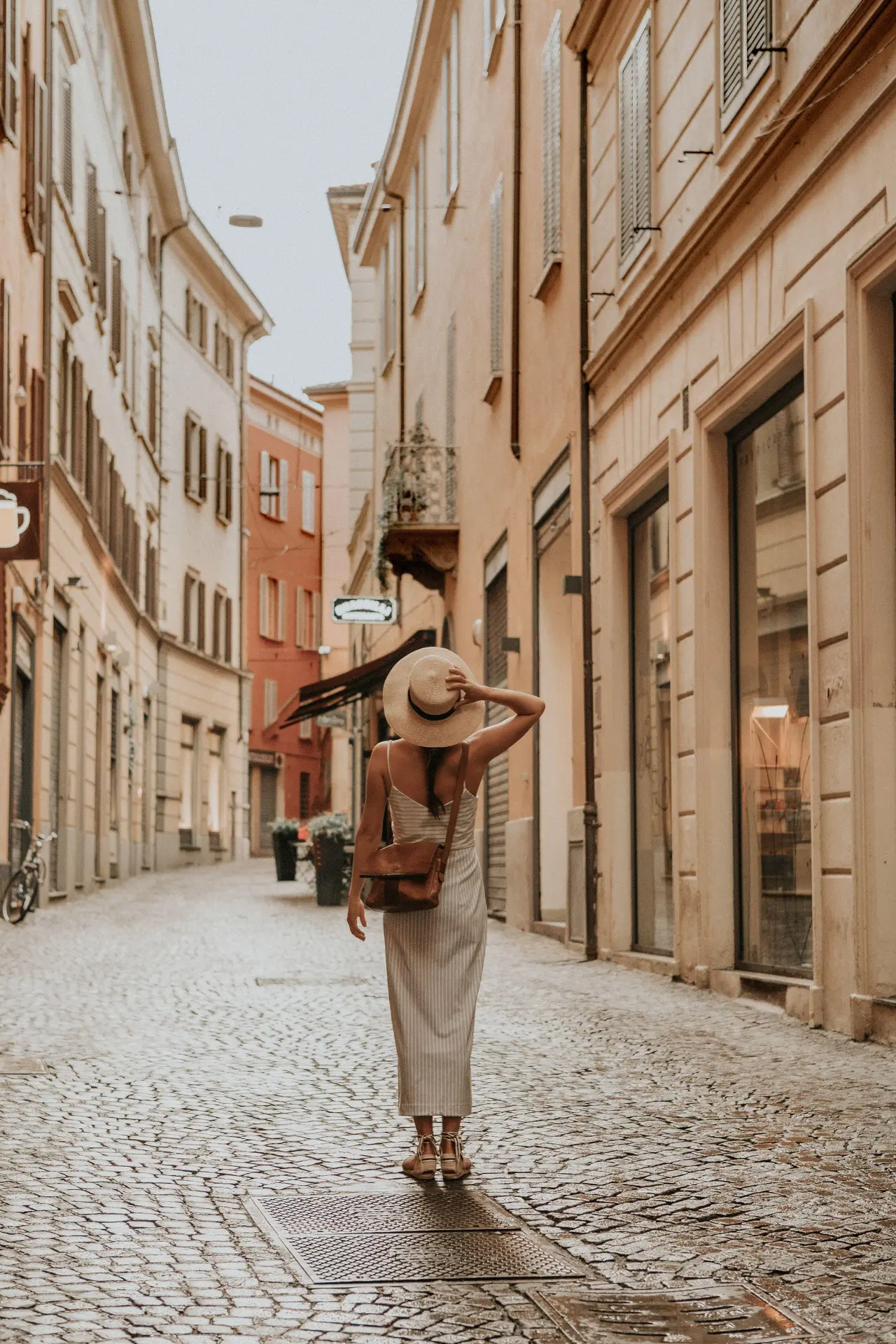 A woman in a wide-brimmed hat walking down a sunlit cobblestone street in Italy